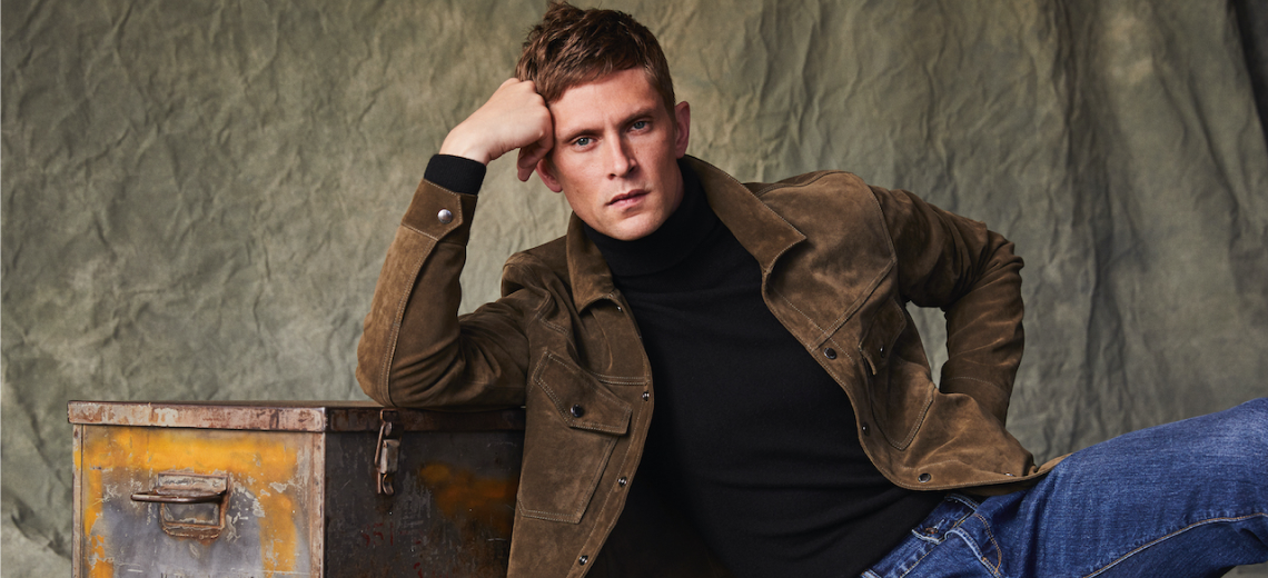 This image shows a male model posing with his head leaned against his hand in a brown jacket, black turtleneck and blue jeans.