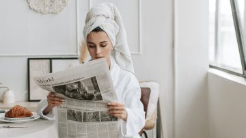 Photograph of a woman sitting a table reading the newspaper in a bathroom and with a towel on her head.