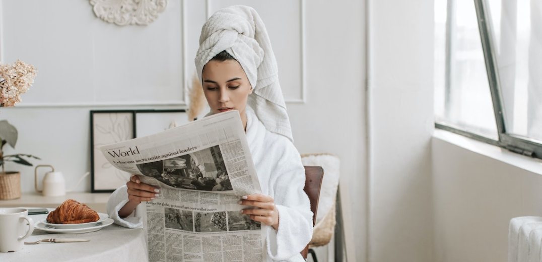 Photograph of a woman sitting a table reading the newspaper in a bathroom and with a towel on her head.